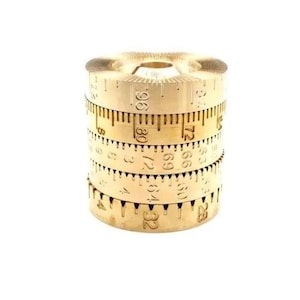 Puede incluir: Una herramienta de medición de metal de color dorado con múltiples anillos giratorios. Cada anillo tiene una escala de medición diferente, incluyendo 28, 32, 64, 69, 72, 80 y 96.
