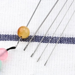 May include: A set of six beading needles with a yellow bead on one of the needles. The needles are laid out on a white fabric with a blue and white pattern.
