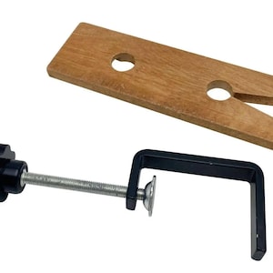 Puede incluir: Un tornillo de banco para carpintería con una base de madera y una abrazadera de metal negro. La base de madera es rectangular con dos agujeros circulares y una muesca en forma de V. La abrazadera tiene una varilla roscada y un pomo negro para apretar.