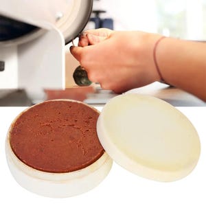May include: A round, white container with a light brown, solid substance inside, next to a white lid. A person's hand is working on a metal object with a grinding wheel in the background.