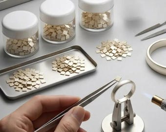 May include: A metal ring held in a silver-colored soldering stand with a torch nearby. Metal tweezers hold a small square piece of metal. Three clear jars with white lids contain many small square metal pieces, and a metal tray holds more pieces.