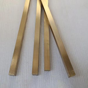 May include: Close-up of four rectangular, golden-colored metal bars. The bars are arranged at different angles, with their flat sides facing the viewer. The metal has a slightly textured surface, reflecting light.