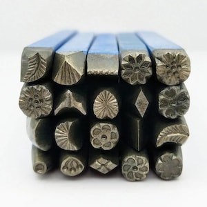 Può includere: Una collezione di timbri metallici con vari disegni decorativi, tra cui floreali, geometrici e a forma di foglia. I timbri hanno manici blu e sono disposti in una formazione impilata, mostrando le dettagliate impressioni che creano.