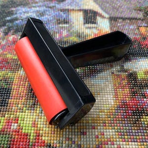 May include: A craft roller tool featuring a smooth, cylindrical red rubber barrel attached to a black plastic frame and handle. The tool is designed for applying pressure to flat surfaces, with a sturdy construction and a T-shaped ergonomic grip.