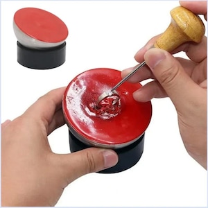 May include: A jewellery setting tool with a ring being worked on. The tool has a wooden handle and a metal tip. The ring is set in a red wax-like substance. Another tool is shown in the background.