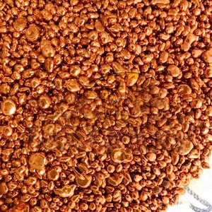 May include: A collection of small, irregular copper metal granules with a shiny, metallic finish. The pieces vary in size and shape, creating a dense, textured surface of reflective, reddish-brown metallic pellets packed closely together.