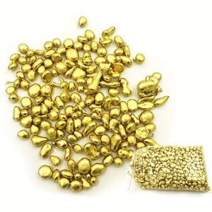May include: A collection of small, irregularly shaped gold nuggets scattered across a white surface, with a clear plastic bag filled with similar nuggets in the lower right corner. The nuggets have a shiny, metallic appearance.