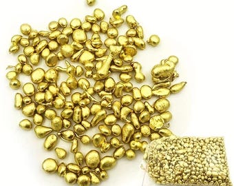 May include: A collection of small, irregularly shaped gold nuggets scattered across a white surface, with a clear plastic bag filled with similar nuggets in the lower right corner. The nuggets have a shiny, metallic appearance.