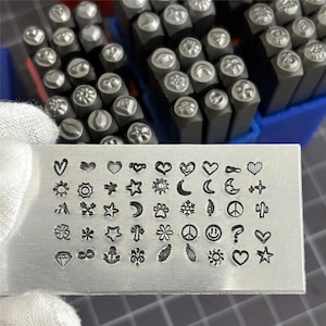 May include: Metal stamping tools featuring a range of design stamps, such as hearts, stars, flowers, and symbols. The stamps are displayed on a metal plate, demonstrating the diverse patterns for crafting and jewellery making.