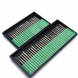 Peut inclure: Deux boîtes en plastique noir remplies de mèches de forage argentées. Chaque boîte contient un insert en mousse verte pour maintenir les mèches en place. Les mèches sont de formes et de tailles variées, adaptées aux travaux de détail.