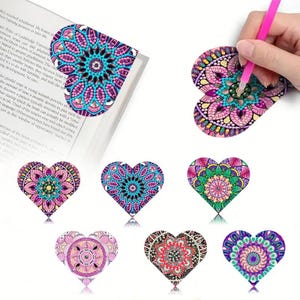 Op de afbeelding: Hartvormige bladwijzers met ingewikkelde mandala-ontwerpen in verschillende kleuren, waaronder roze, blauw en groen. De bladwijzers liggen op een boek en een hand houdt een roze potlood vast. De bladwijzers zijn een decoratief item.