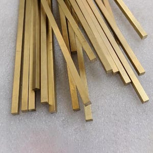 Pode incluir: Uma coleção de barras retangulares de latão, cada uma com um tom dourado. As barras são de comprimentos variados e estão dispostas em um padrão disperso. As barras de metal são provavelmente para artesanato ou uso industrial.