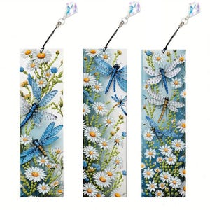 May include: Three rectangular bookmarks with dragonfly and daisy designs. Each bookmark features a different dragonfly and daisy arrangement, with a blue and white colour scheme. Each bookmark has a black string and a crystal charm attached at the top.