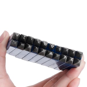 May include: A set of fourteen rectangular metal stamping tools arranged in two rows. Each tool features a distinct engraved symbol on its end, including zodiac signs, a heart, a smiley face, a four-leaf clover, and a musical treble clef.