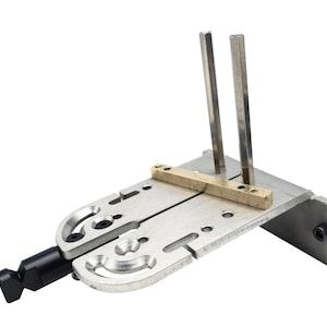 May include: A metal jig tool featuring a silver-toned base plate with curved slots and circular holes, a horizontal brass-coloured bar, two vertical silver rods, and a black adjustment knob at the end, designed for precise alignment and positioning.