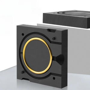 以下が含まれることがあります： 中央に金色の金属製リングインサートを備えた円形の空洞を持つ、黒い正方形の工業用金型コンポーネント。このブロックには、小さな円筒形の合わせピン、漏斗状の側面開口部、および一致する黒い上部プレートセクションが含まれています。