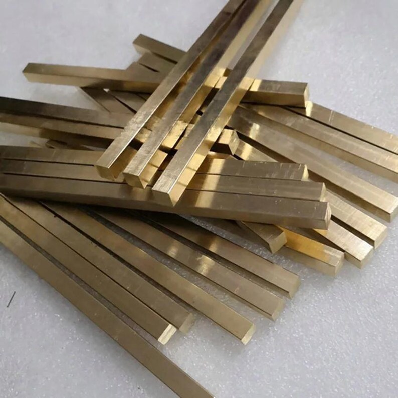 May include: A collection of rectangular brass bars, each with a polished, golden finish. The bars are arranged in a scattered pile, with their edges and surfaces reflecting light. The overall composition suggests a supply of raw materials or components.