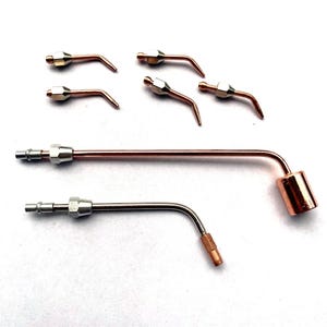 Op de afbeelding: Een set van zeven metalen las- of soldeertips en mondstukken. De set bevat vijf kleine, schuine koperkleurige tips met zilverkleurige connectoren en twee langere, gebogen metalen buizen, waarvan één met een koperkleurige cilindrische kop.