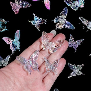 Puede incluir: Una colección de pequeños adornos translúcidos con forma de mariposa y acabado iridiscente. Estas piezas decorativas presentan delicados patrones en las alas y están esparcidas sobre una superficie oscura, con varias sostenidas en una palma.