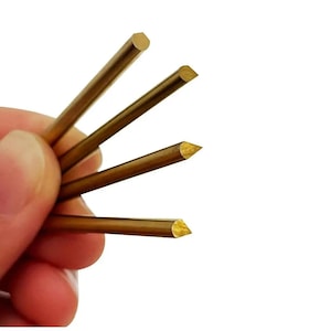 May include: Close-up of several small, golden-colored metal rods. Some rods have a rounded end, while others are cut at an angle, creating a pointed tip. The rods are held in a hand against a white background.