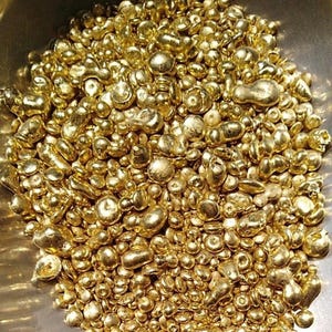 May include: A collection of small, irregular, metallic gold-coloured nuggets or casting grains. Each piece features a smooth, rounded, and reflective surface with varying shapes and sizes, creating a dense, textured pile of shiny, bright yellow metal particles.