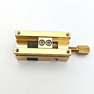 May include: A small, gold-colored vise with a threaded adjustment screw and a knurled knob. The vise has two parallel jaws and is secured with small screws. The object is designed to hold small items.
