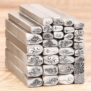 Könnte beinhalten: Ein Stapel Metallstempel mit Wolkenmotiven. Die Stempel sind rechteckig mit strukturierten Seiten und verschiedenen Wolkenmustern, die in die Schlagfläche geschnitzt sind. Die Stempel sind silberfarben.