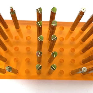 May include: An orange acrylic display stand filled with numerous brown and gold-tipped tools. The tools are arranged vertically, with some featuring green and white accents. The stand has a rectangular base with evenly spaced circular cutouts.