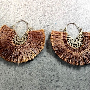 May include: A pair of fan-shaped earrings featuring brown fringe tassels attached to a gold-toned metal filigree semi-circle frame. The earrings include a thin gold-toned wire hoop closure at the top, designed to hang from the earlobe.