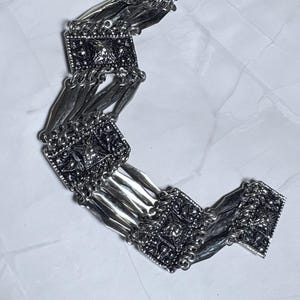 Könnte beinhalten: Ein silberfarbenes Armband mit vier quadratischen, texturierten Metallplatten, die durch mehrere dünne, geschwungene Silberstäbe verbunden sind. Die quadratischen Platten haben ein detailliertes Prägemuster mit dunkler, oxidierter Oberfläche.