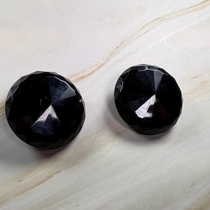 May include: Two round, black buttons with a faceted, diamond-like surface texture. Each button features a central point with multiple triangular facets radiating outward toward the circular edge, creating a reflective and geometric appearance.
