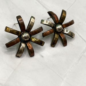 May include: A pair of stud earrings shaped like flowers with eight petals each. The petals alternate between a metallic copper tone and a tarnished silver tone. A small, smooth, polished silver-colored sphere is centered in the middle of each flower.