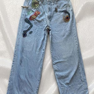 May include: Light blue denim jeans featuring a wide-leg silhouette and a raw, frayed hem. The front of the jeans is decorated with various colourful, textured fabric patches and embroidered shapes, including abstract forms and a small fern leaf.