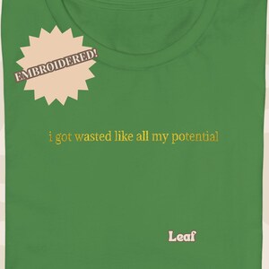 May include: Green t-shirt with the embroidered text "i got wasted like all my potential" in gold. A tan starburst graphic with the word "EMBROIDERED!" is on the upper left. The word "Leaf" is embroidered at the bottom.