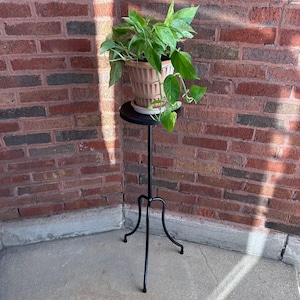 May include: A potted plant with green and yellow leaves sits on a black, round plant stand. The stand has a slender, black metal stem and three curved legs. The plant pot is a light brown color and is set against a red brick wall.