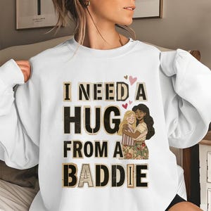 Peut inclure: Sweat-shirt blanc avec l'inscription "I NEED A HUG FROM A BADDIE" dans une police vieillie. Le motif comprend deux femmes qui s'embrassent, avec de petits cœurs roses au-dessus. Le sweat-shirt est de style décontracté et confortable.