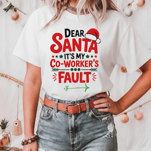 May include: White t-shirt with the text "Dear Santa It's My Co-Worker's Fault" in red and green. The design includes a Santa hat and arrow. The shirt is paired with denim shorts and a brown belt.