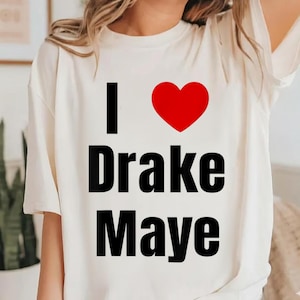 Może przedstawiać: Kremowa koszulka z napisem "I ❤️ Drake Maye" czarnymi literami i czerwonym sercem. Koszulka jest w stylu casual, odpowiednia do noszenia na co dzień.