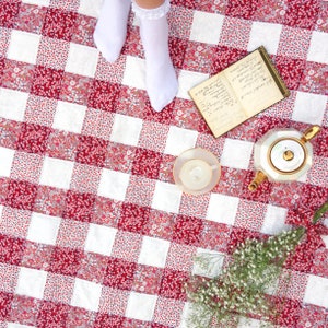 May include: A red and white checkered picnic blanket with a teapot, a teacup and saucer, a book, and white flowers.