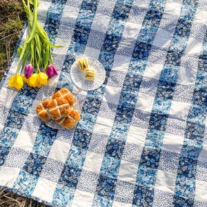Puede incluir: Una manta de picnic a cuadros blancos y azules con un patrón floral. La manta está extendida sobre la hierba y tiene un plato de pasteles y un ramo de flores amarillas y moradas.
