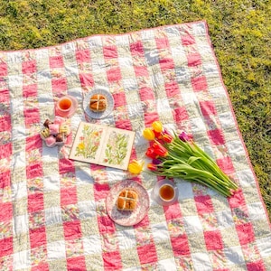 May include: A picnic setting on a sunny day, featuring a pink and white checkered quilt on grass. The quilt is set with tea cups, plates with pastries, a book with floral illustrations, and a bouquet of colorful tulips.