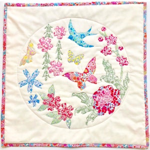 Può includere: Un quadrato di tessuto bianco con un motivo floreale rosa e blu. Il motivo è in un cerchio e presenta farfalle, fiori e uccelli. Il tessuto è trapuntato e ha un bordo rosa e blu.