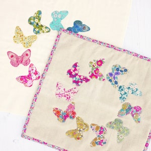 Pode incluir: Dois quadrados de tecido branco sujo decorados com aplicações de borboletas coloridas. Um quadrado tem borboletas dispostas em círculo, o outro tem borboletas espalhadas pela superfície. As borboletas apresentam padrões florais em rosa, azul, amarelo e verde.