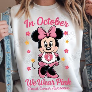 May include: White sweatshirt with a pink Minnie Mouse graphic holding a pink heart with a ribbon. The text "In October We Wear Pink Breast Cancer Awareness" is printed on the shirt. Pink and yellow flowers are scattered around the graphic.