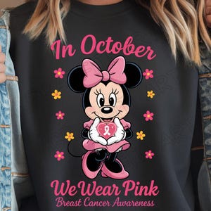 May include: Black sweatshirt featuring Minnie Mouse in pink attire, holding a pink heart with a ribbon. The text "In October We Wear Pink Breast Cancer Awareness" is displayed in pink.