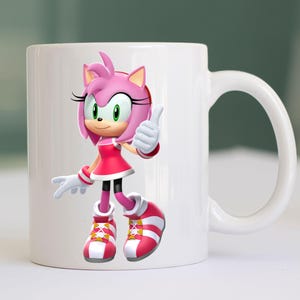 May include: White ceramic mug featuring a colorful illustration of Amy Rose from the Sonic the Hedgehog series. Amy Rose is depicted in her signature pink dress, shoes, and quills, giving a thumbs-up gesture. The mug has a curved handle.