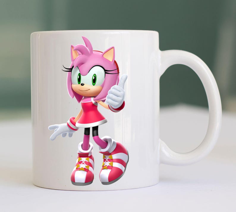 May include: White ceramic mug featuring a colorful illustration of Amy Rose from the Sonic the Hedgehog series. Amy Rose is depicted in her signature pink dress, shoes, and quills, giving a thumbs-up gesture. The mug has a curved handle.