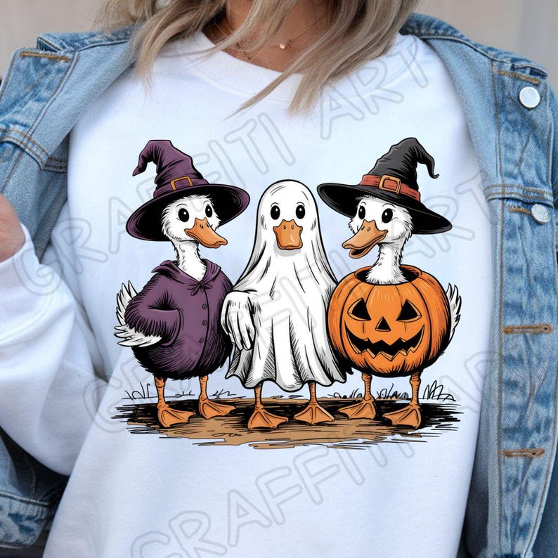 May include: A white graphic tee featuring three ducks dressed for Halloween. One duck wears a witch hat and purple cloak, another is a ghost, and the third is inside a carved jack-o'-lantern. The ducks stand on a patch of brown grass.