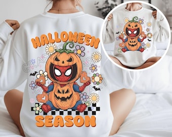 May include: White sweatshirt featuring a Halloween-themed graphic. The design includes a pumpkin-Spider-Man character, flowers, and the words "HALLOWEEN SEASON" in orange. The sweatshirt has a crew neck and long sleeves.