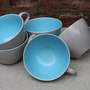 May include: A set of grey ceramic cups with a light blue interior. The cups have a simple, modern design with a curved handle. The cups are stacked and arranged on a neutral-coloured surface.